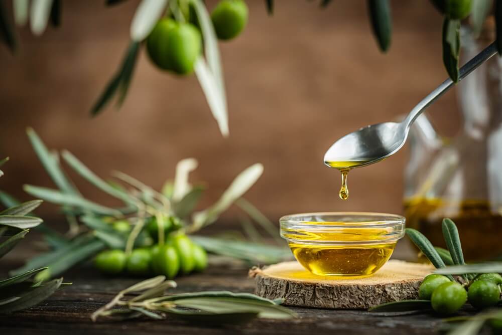 Skledica oljčnega olja na deščici, obdana z vejami oljke, nad skledico žlica, s katere pada kapljica olja